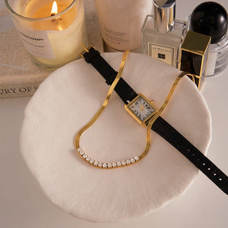 Gold watch with black strap on a white stand, surrounded by candles and bottles.