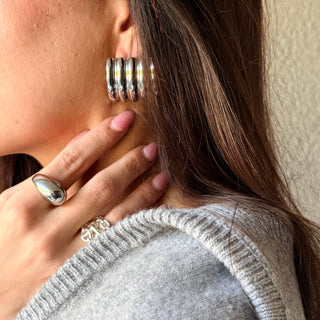 A close-up lifestyle shot of a woman wearing a chunky, multi-layered silver hoop earring and a matching silver ring on her finger, wearing a light gray knit sweater.