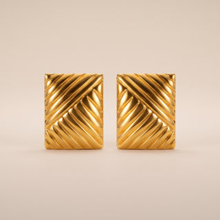 A pair of gold square cufflinks with a textured diagonal chevron pattern, displayed on a neutral beige background.