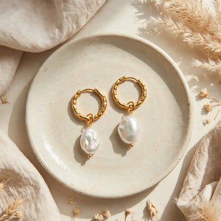 A pair of gold hoop earrings with irregular white baroque pearl drops, displayed on a round ceramic plate surrounded by beige linen fabric and dried pampas grass.