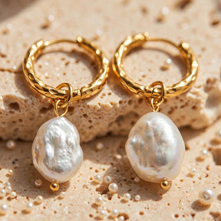 A pair of gold hoop earrings with large, irregular white baroque pearl drops, displayed on a textured beige stone surface with scattered small pearls.