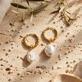 A pair of gold hoop earrings with dangling irregular white baroque pearls, styled on a textured beige stone surface with linen fabric, dried grass, and greenery.