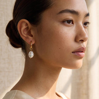 A close-up side profile of a woman wearing a gold dangle earring with a white teardrop-shaped pearl, set against a soft beige textured background.