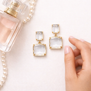 Gold earrings with clear stones on a white surface with a perfume bottle and pearls.