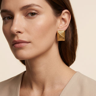 A close-up of a woman wearing a textured gold geometric stud earring, shown from the chest up against a warm beige background.