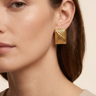 A close-up of a woman wearing a textured gold geometric stud earring, shown from the chest up against a warm beige background.