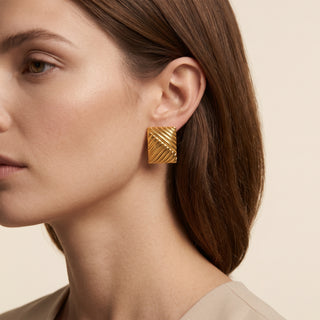 A close-up of a woman wearing a textured gold square stud earring with diagonal ridges, set against a soft beige background.