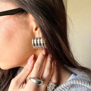 A close-up lifestyle shot of a woman wearing a chunky silver hoop earring with multiple stacked horizontal bands. She is wearing a gray knit sweater, a silver dome ring, and a silver floral ring, with dark hair and black-framed glasses visible.