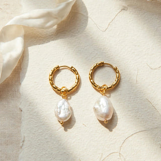 A pair of gold hoop earrings with dangling baroque pearl drops, displayed on a textured beige surface with a cream ribbon.