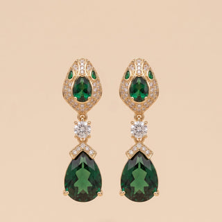 A pair of gold drop earrings featuring large green teardrop gemstones, smaller green stones at the top, and clear accent stones, displayed on a solid beige background.