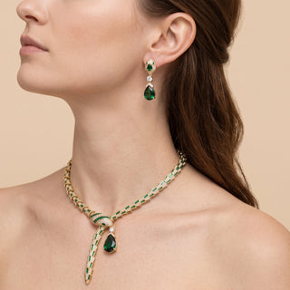 A close-up of a woman wearing a gold and emerald green jewelry set, featuring a braided necklace with a teardrop pendant and a matching drop earring, against a solid beige background.