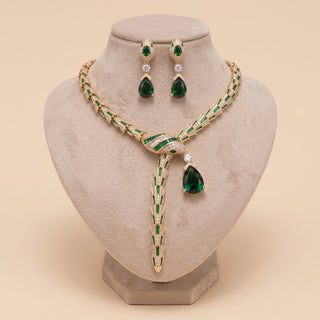 Gold and emerald green jewelry set displayed on a beige velvet jewelry bust, including a necklace with a long teardrop pendant and matching drop earrings with clear stones.
