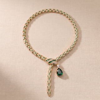 A luxury gold and emerald green woven chain necklace with a knot closure and a teardrop-shaped dark green gemstone pendant, displayed on a textured beige fabric background.