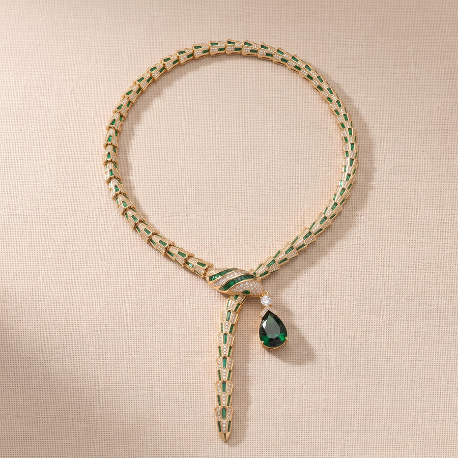 A luxury gold and emerald green woven chain necklace with a knot closure and a teardrop-shaped dark green gemstone pendant, displayed on a textured beige fabric background.