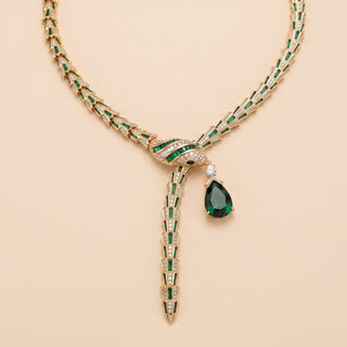 A luxury gold and emerald green necklace with a teardrop-shaped emerald pendant and diamond accents, displayed on a solid beige background.
