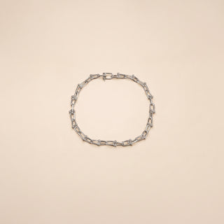 A silver-toned chain link bracelet with elongated oval links and small geometric accents, arranged in a circle on a solid pale beige background.