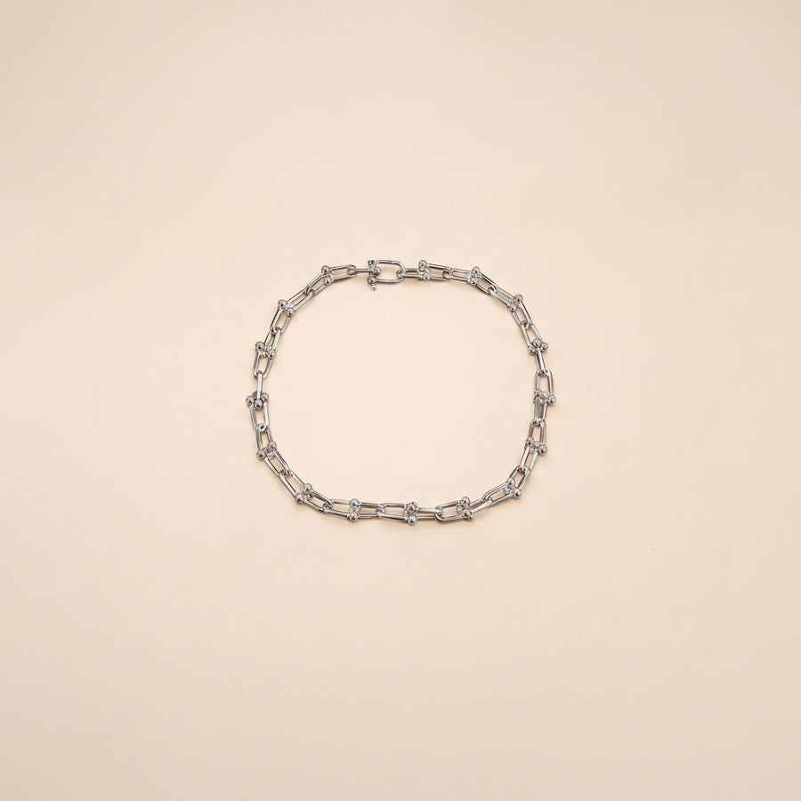 A silver-toned chain link bracelet with elongated oval links and small geometric accents, arranged in a circle on a solid pale beige background.