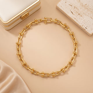 A gold chain necklace with a repeating ball-and-link design, arranged in a circle on a warm beige surface with a cream jewelry box and a stone slab as styled props.