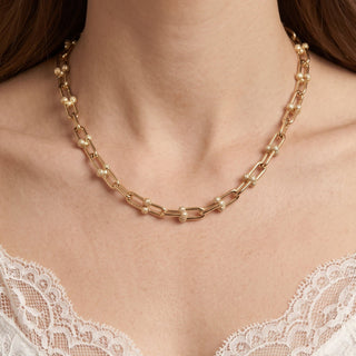 A close-up of a model wearing a silver-toned link necklace with small clear gemstone accents, shown against the model's neck and shoulders.