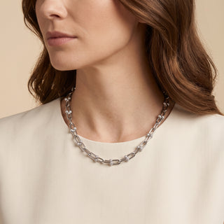 A close-up of a woman wearing a chunky silver chain-link necklace with clear crystal-like beads, paired with a cream-colored top against a warm beige background.