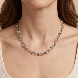 A close-up of a model wearing a silver-toned link necklace with small clear gemstone accents, shown against the model's neck and shoulders.