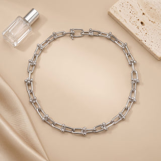A silver chain necklace with elongated links and small bead details, arranged in a circle on a beige satin fabric. A small clear perfume bottle and a textured stone slab are styled props in the background.