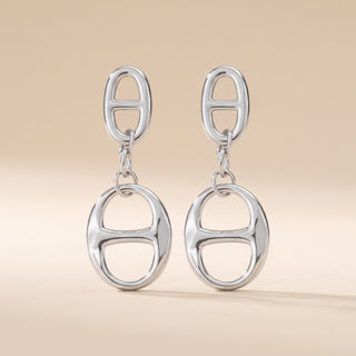 A pair of silver drop earrings featuring a small oval link at the top and a larger oval link dangle below, set against a solid beige background.