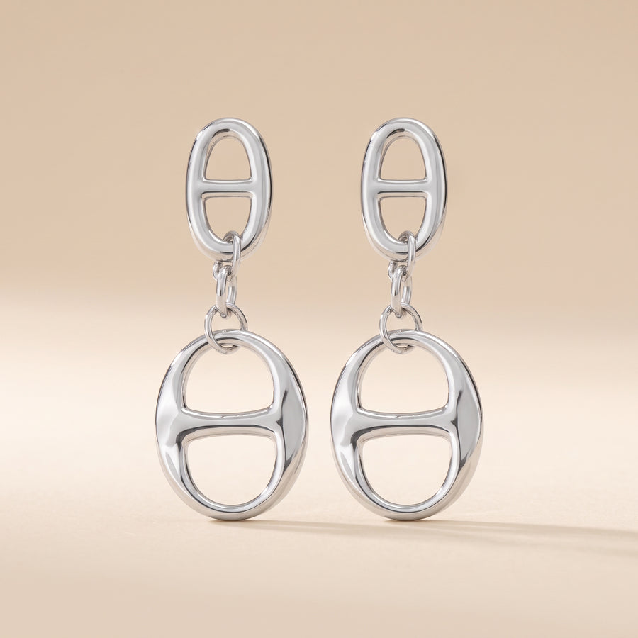 A pair of silver drop earrings featuring a small oval link at the top and a larger oval link dangle below, set against a solid beige background.