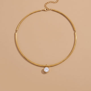 A gold-toned bangle-style necklace with a single clear round gemstone pendant, displayed on a warm beige background.