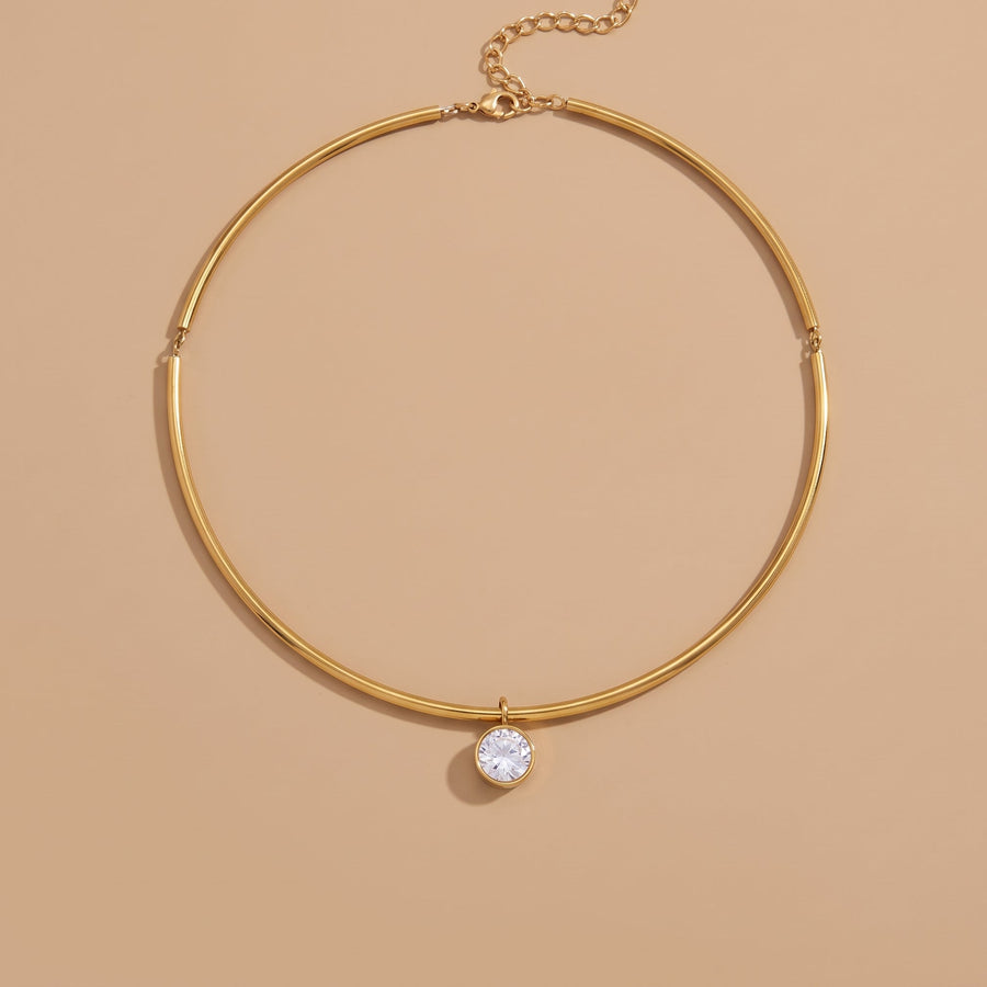 A gold-toned bangle-style necklace with a single clear round gemstone pendant, displayed on a warm beige background.