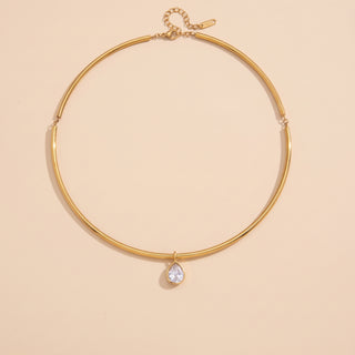A gold-toned thin choker necklace with a small teardrop-shaped clear gemstone pendant, displayed on a solid pale peach background.